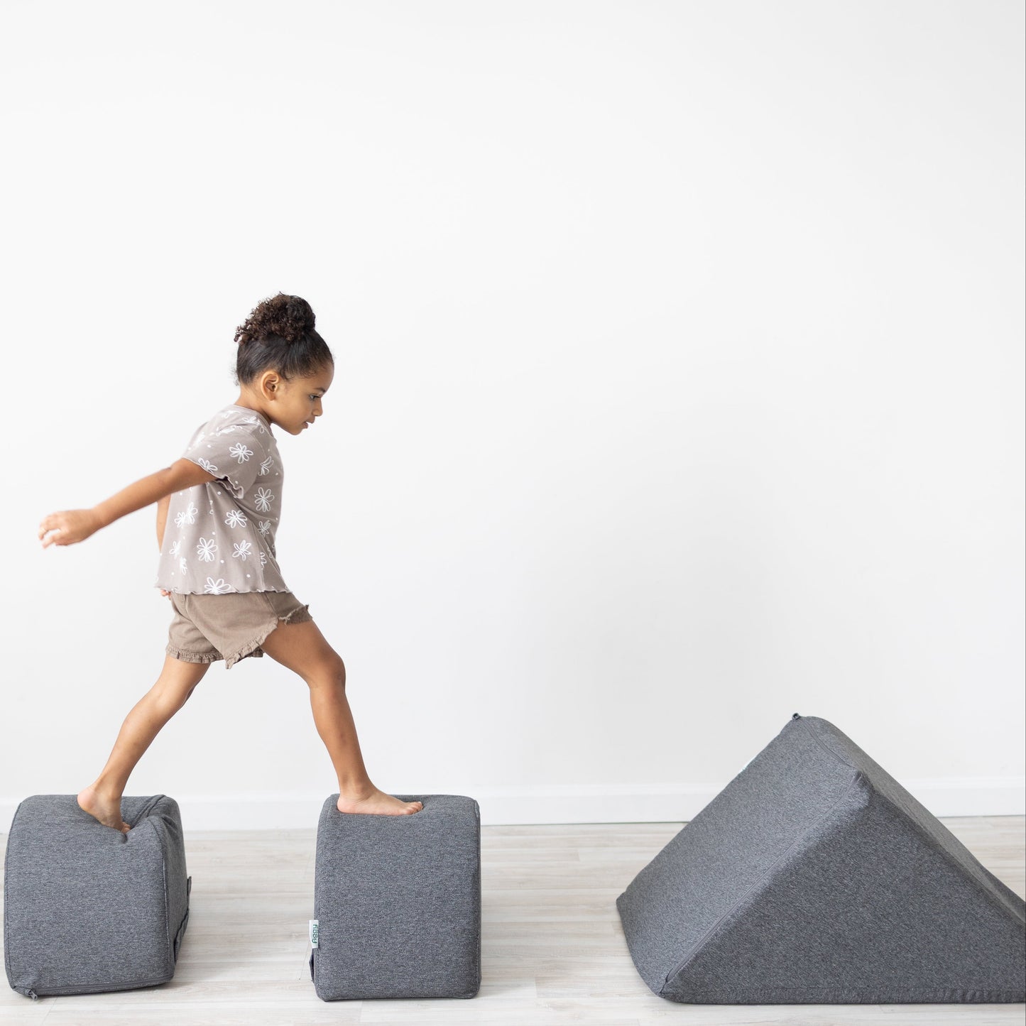 Child playing on gray foam blocks in a minimalistic room with a white wall and wooden floor.