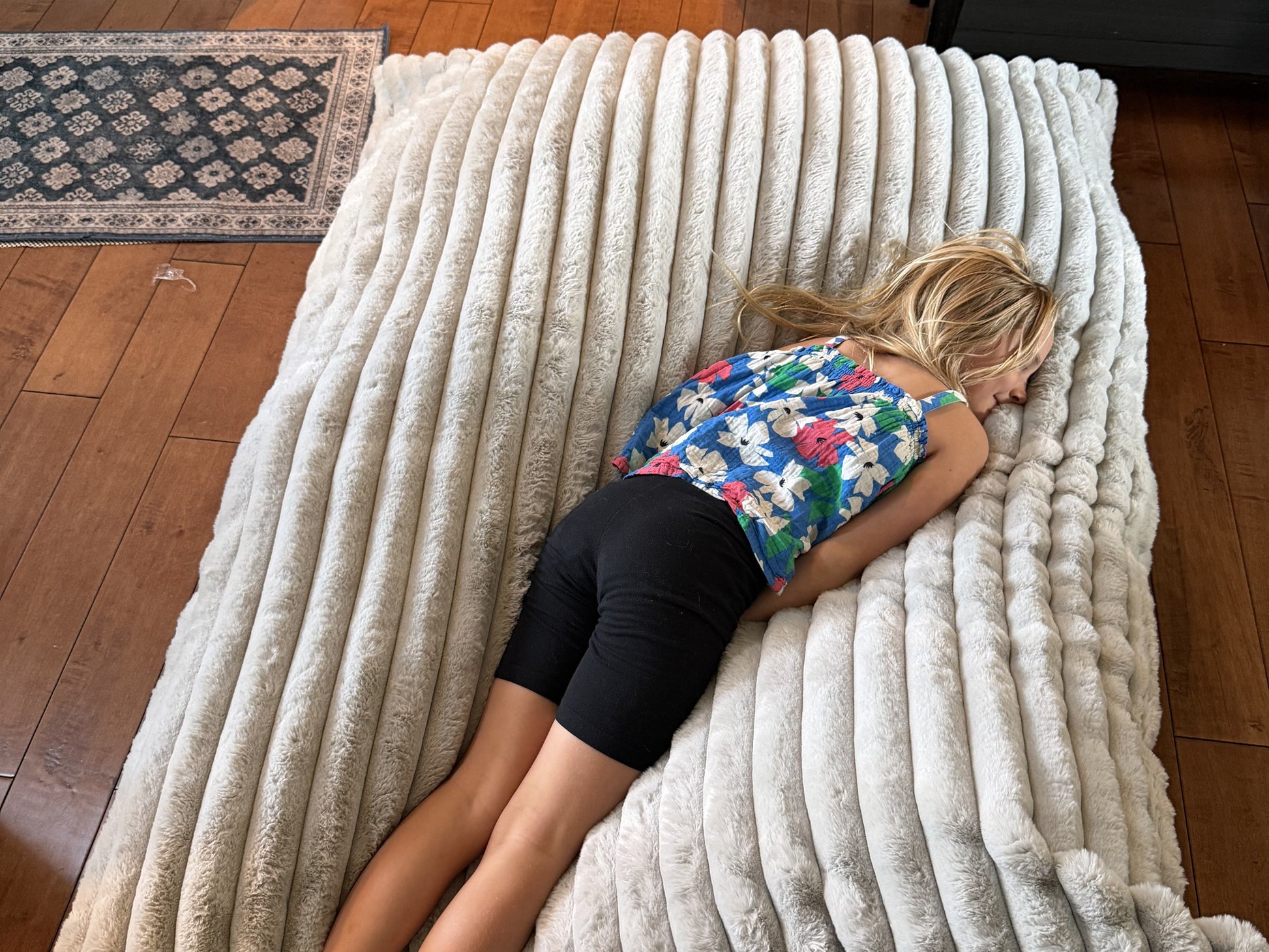 A child resting on The Baggy faux fur crash pad, shown flat on a wood floor
