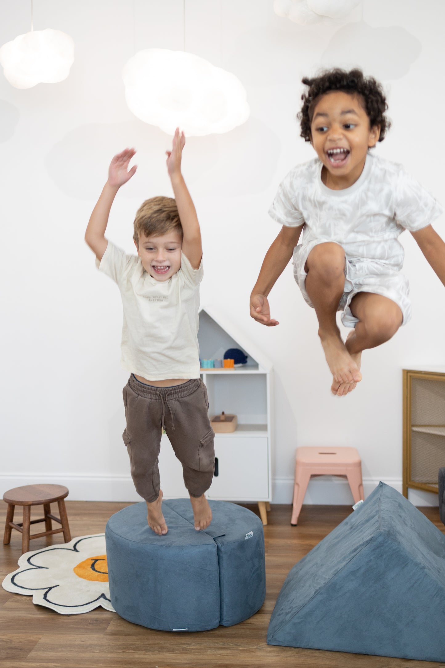 Two children jumping on a blue ottoman in a room with toys and furniture.
